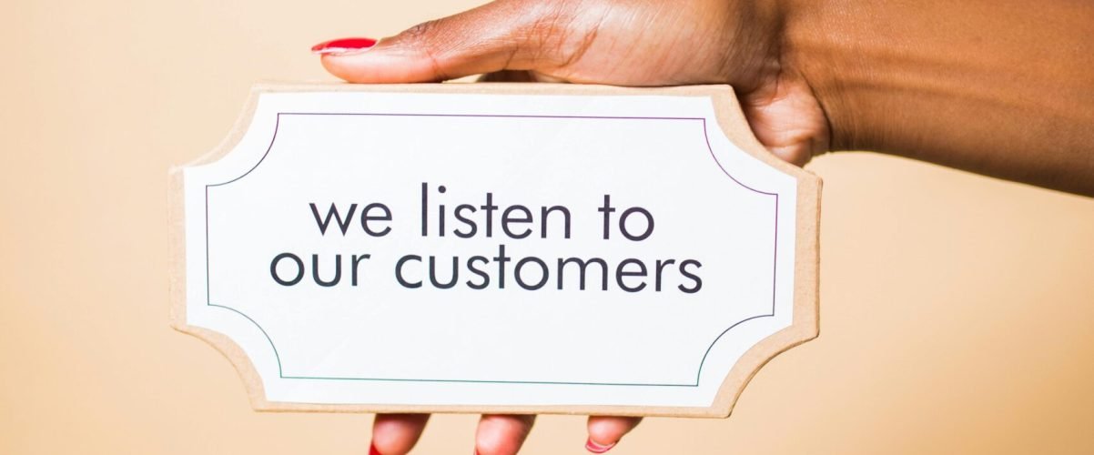 A hand holding a sign saying 'we listen to our customers' on a neutral background.
